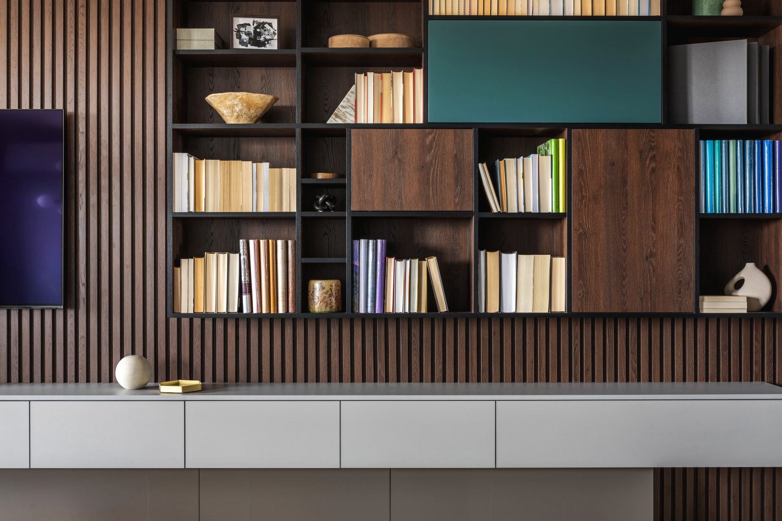 Modern bookcase in lamells wall. Grey chest of drawers. Bookcase a lot of books and accessories. Herringbone wooden parquet. Cat in open spaces.