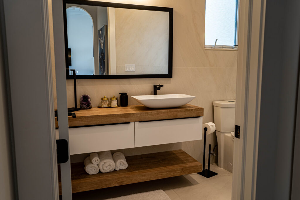 Modern custom bathroom vanity with wood countertop and open shelf, white vessel sink, and black framed mirror in a minimalist design. Matching dual sink bathroom setup with open shelf storage and mirror lighting.