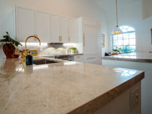 Traditional kitchen with granite countertops, white cabinetry, and gold fixtures, lit by large windows.