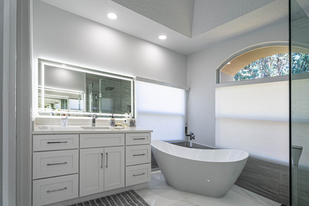 Custom white bathroom vanity with shaker-style cabinets and integrated double sink, paired with a modern freestanding bathtub in a bright, contemporary bathroom. Freestanding white tub beside a vanity with LED mirror and gray flooring.